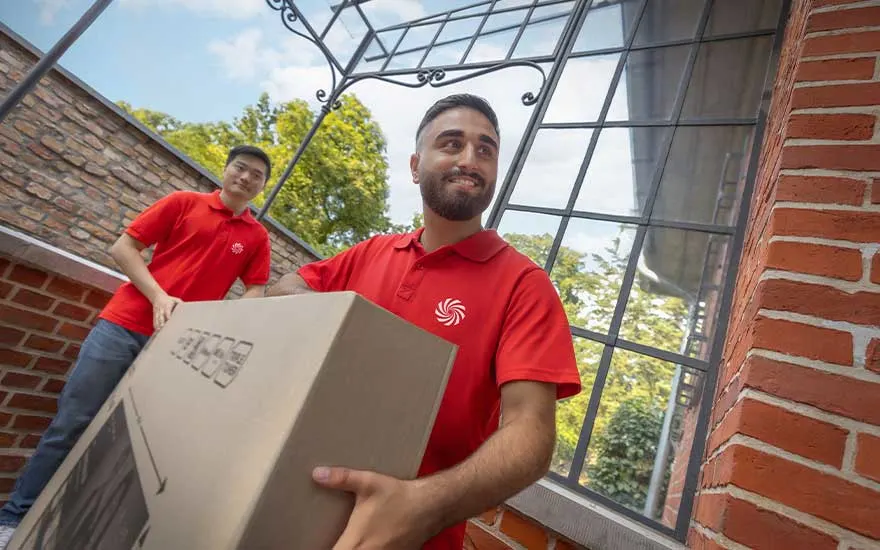Twee koeriers in rode polo’s dragen samen een groot pakket bij een huis met bakstenen muren en glasramen, klaar voor levering.