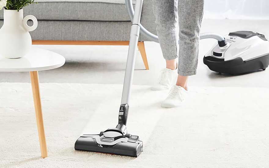 A person vacuums a white carpet in a stylish room with a canister hoover, the floor looks fresh and clean. 