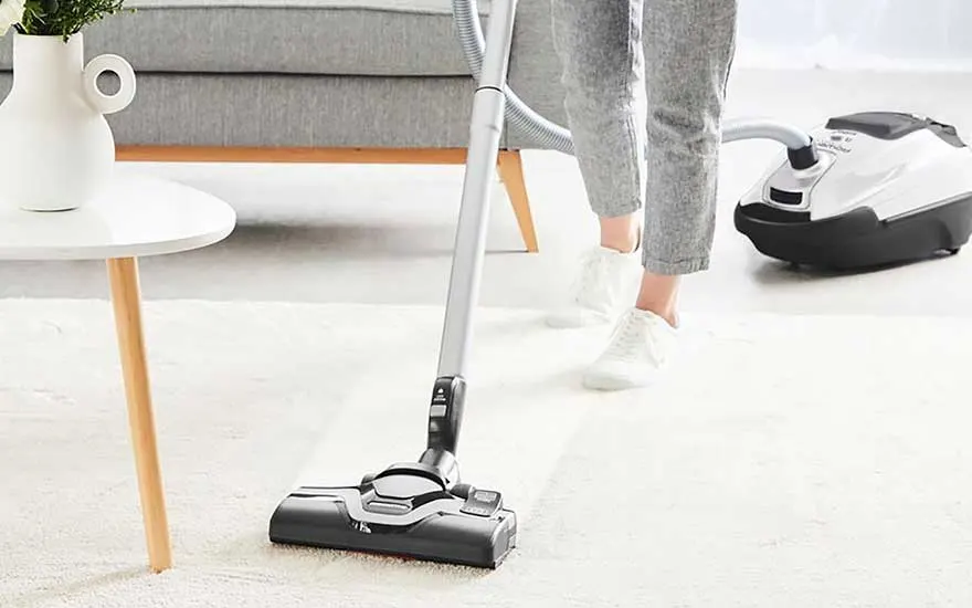 A person vacuums a white carpet in a stylish room with a canister hoover, the floor looks fresh and clean.