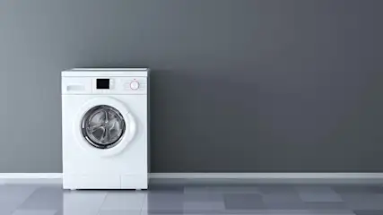 Een witte wasmachine staat tegen een grijze muur op een grijze tegelvloer.