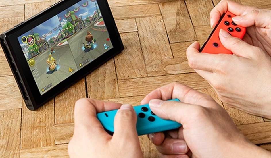 Twee mensen spelen Mario Kart op een Nintendo Switch met rode en blauwe Joy-Con controllers op een houten tafel.