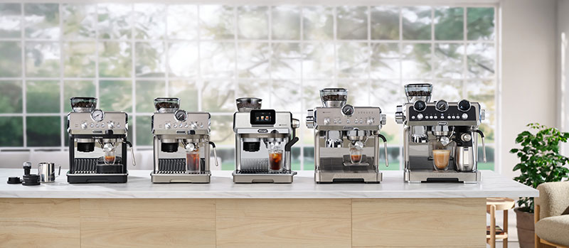 Row of five espresso machines on a kitchen island in front of a large window, each with a coffee cup beneath the group head.