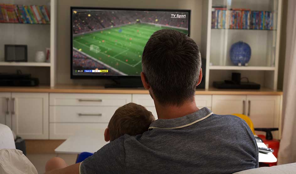 An adult sits with a child on their lap, watching a football match on television in a cozy living room.