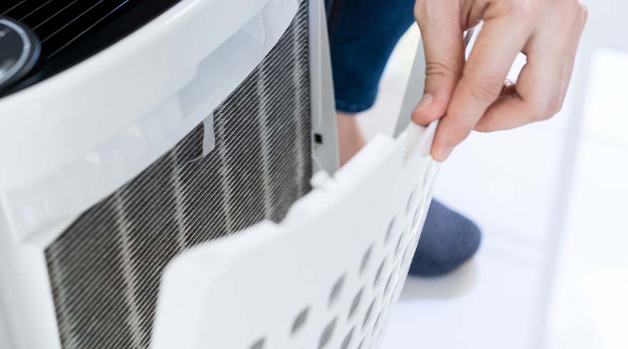 Een hand verwijdert een wit filter met gaten van een wit apparaat met grijze roosters. Blauwe kleding is zichtbaar.