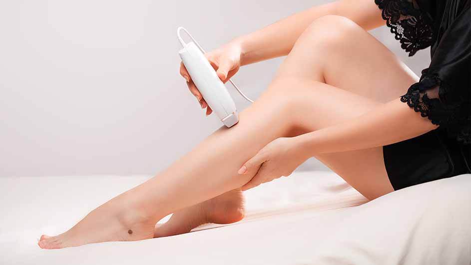 Een vrouw gebruikt een wit apparaat met snoer op haar been. Ze draagt een zwarte kamerjas met kant.