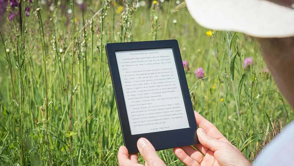 Een persoon houdt een e-reader vast in een veld met groen gras en bloemen. Het scherm van de e-reader toont tekst.