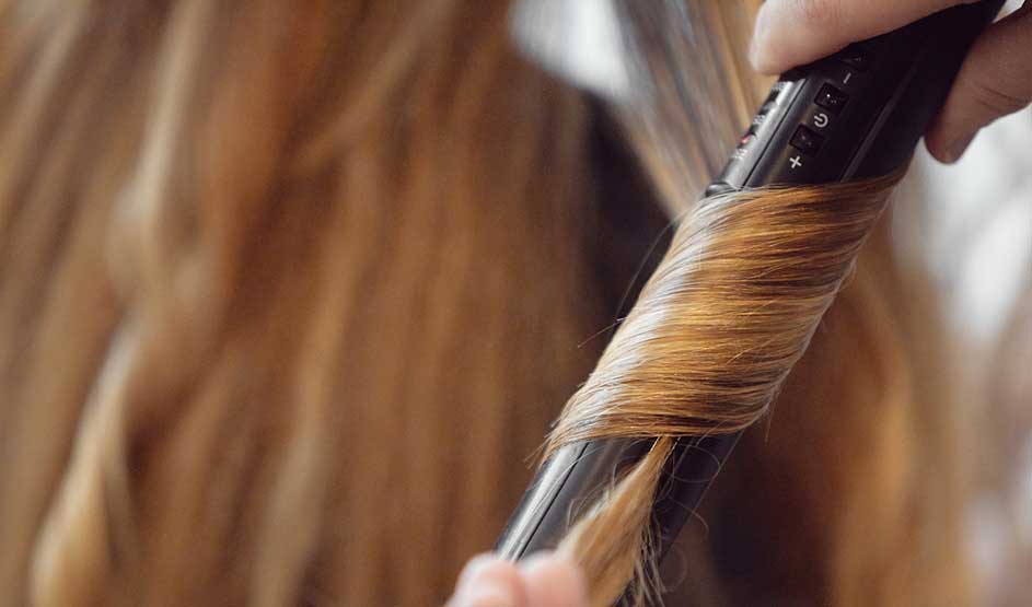A strand of light brown hair is wrapped tightly round a curling wand, while a hand carefully shapes the style.