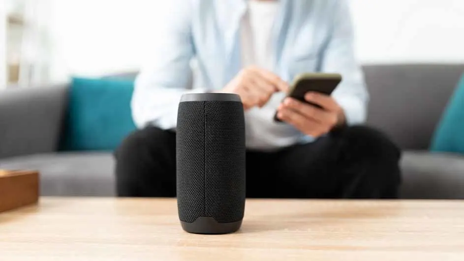 Een zwarte speaker staat op een houten tafel, met op de achtergrond een persoon met een telefoon op een grijze bank.