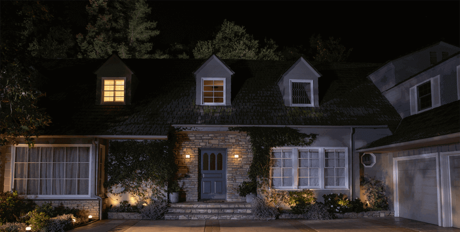 Een huis met een donker dak en stenen details, verlicht door buitenlampen en ramen. Er zijn bomen op de achtergrond.