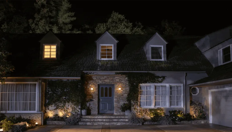 Een huis met een donker dak en stenen details, verlicht door buitenlampen en ramen. Er zijn bomen op de achtergrond.