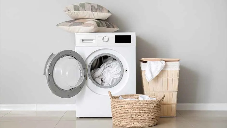 A white washing machine with its door open contains laundry. A basket and a laundry hamper are beside it, with cushions stacked on top. No brand visible.