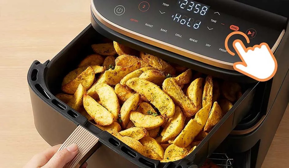 A hand slides the drawer full of seasoned potato wedges into a black Ninja air fryer with digital display and illuminated buttons.