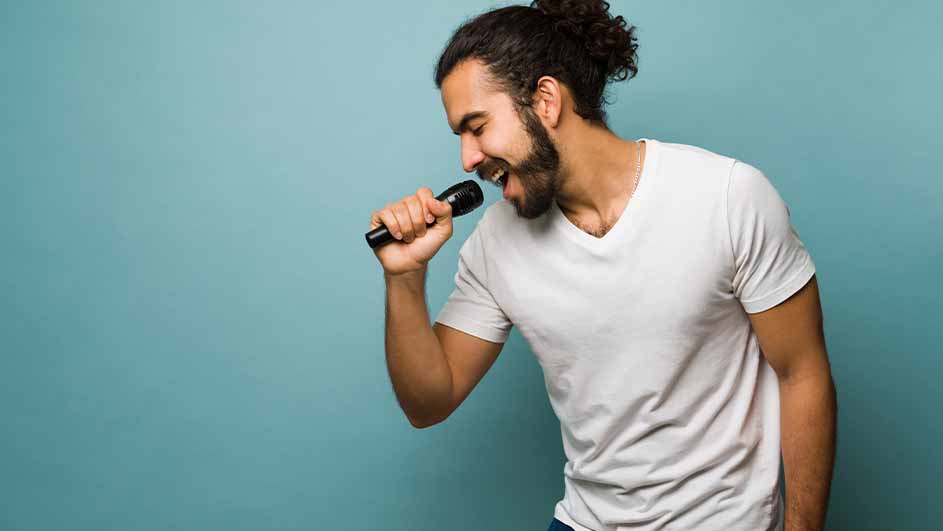 Een man met een baard en opgestoken haar zingt in een microfoon tegen een blauwe achtergrond, gekleed in een wit T-shirt.