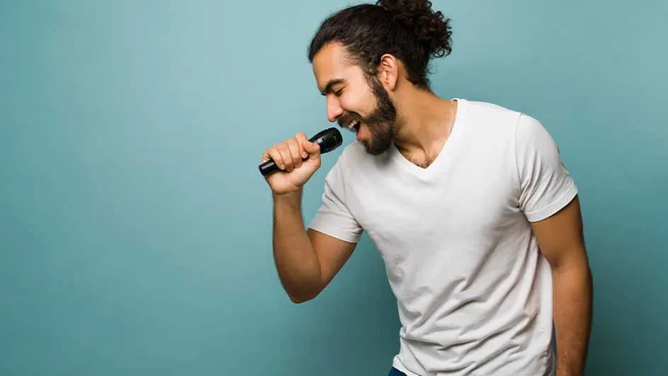Een man met een baard en opgestoken haar zingt in een microfoon tegen een blauwe achtergrond, gekleed in een wit T-shirt.