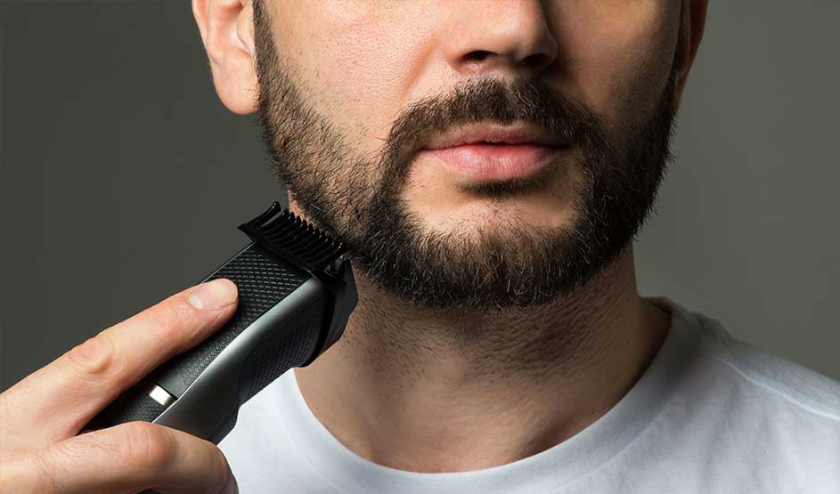 Een man trimt zijn baard met een zwarte tondeuse langs de kaak; alleen zijn ondergezicht en witte shirt zijn zichtbaar.
