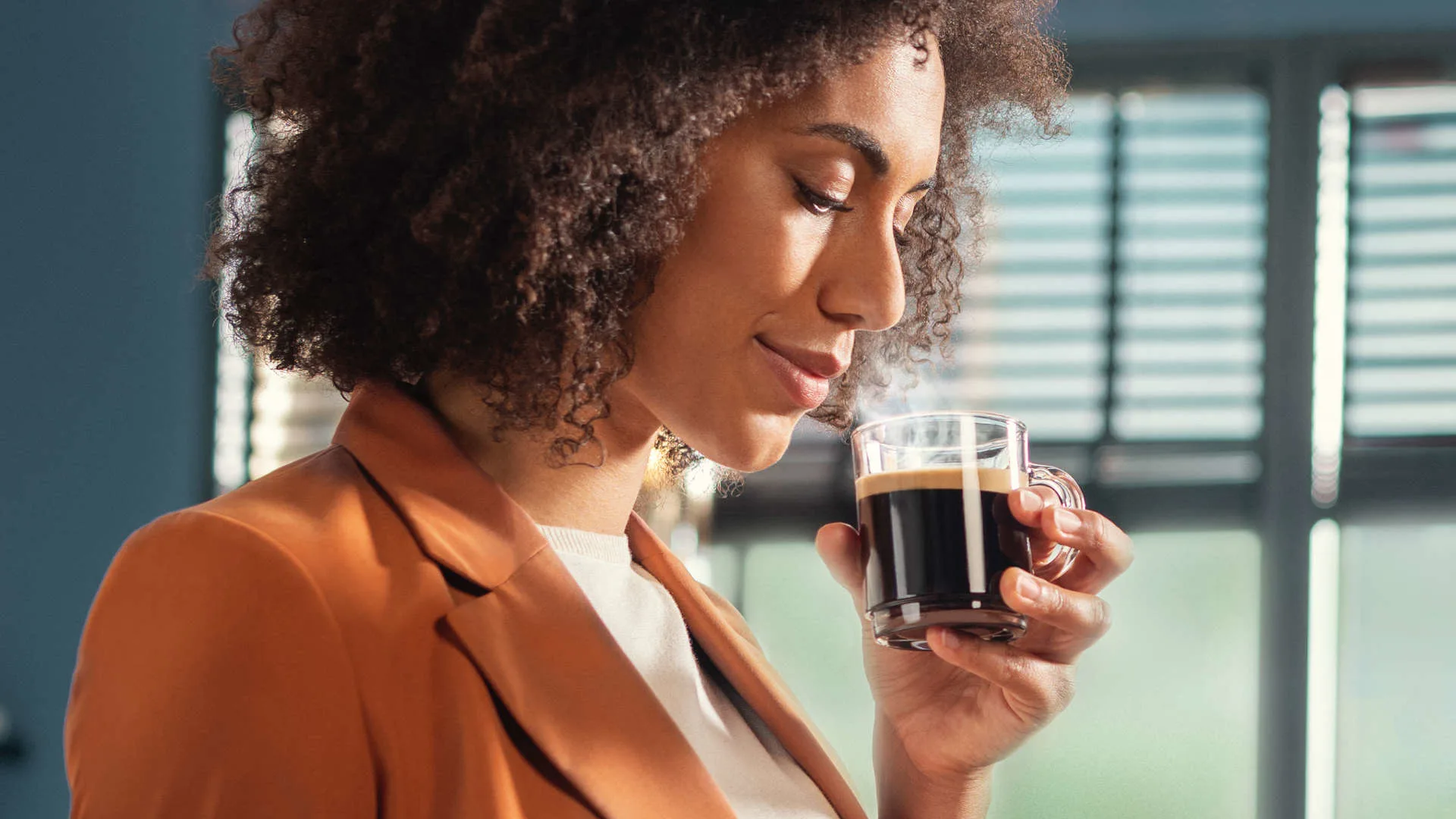 Een vrouw met een oranje jas houdt een glas zwarte koffie vast. Op de achtergrond zijn jaloezieën te zien.