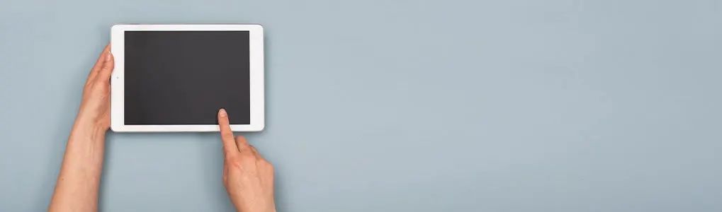 Two hands hold a white tablet with a black screen on a gray background; one finger touches the screen.