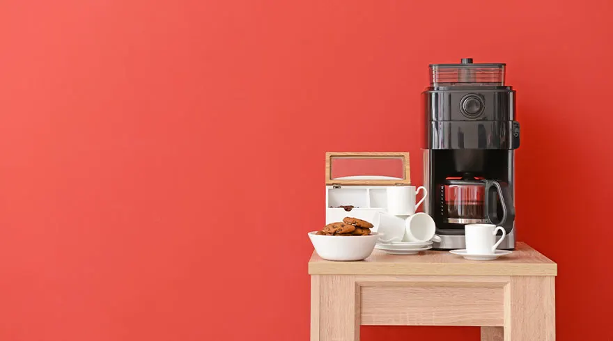On a table against a red wall are a coffee maker, a bowl of cookies, and several white cups and saucers.