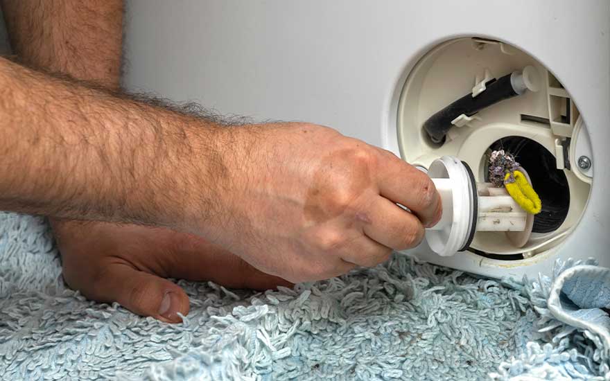 I see someone cleaning a washing machine filter. The person is holding the filter, ready to clean it. There's visible dirt and debris inside the machine. The person is sat on a soft, blue bath mat.