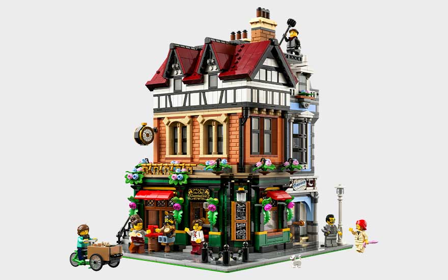 Gedetailleerd LEGO-hoekgebouw met rode daken, winkel op straatniveau, bloemen, balkon en minifiguren rondom.