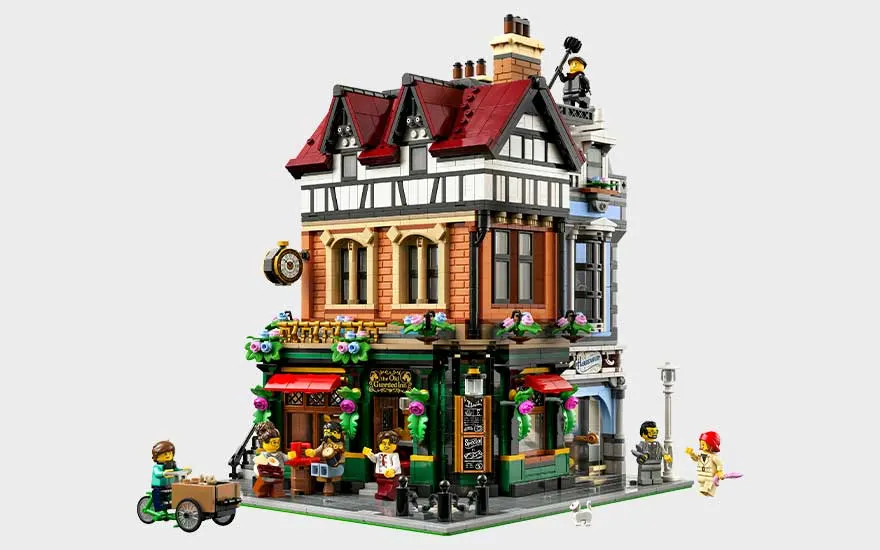 Gedetailleerd LEGO-hoekgebouw met rode daken, winkel op straatniveau, bloemen, balkon en minifiguren rondom.