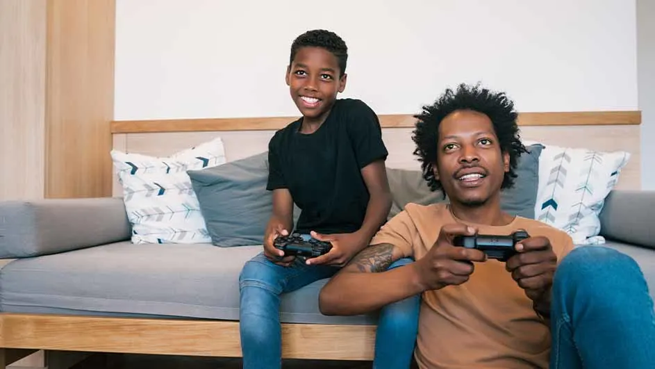 Twee mannen zitten op een grijze bank met kussens en spelen videospelletjes met zwarte controllers.