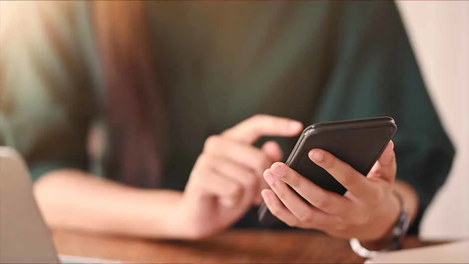 Een persoon in een groen shirt gebruikt een zwarte smartphone aan een houten tafel, met een laptop links.