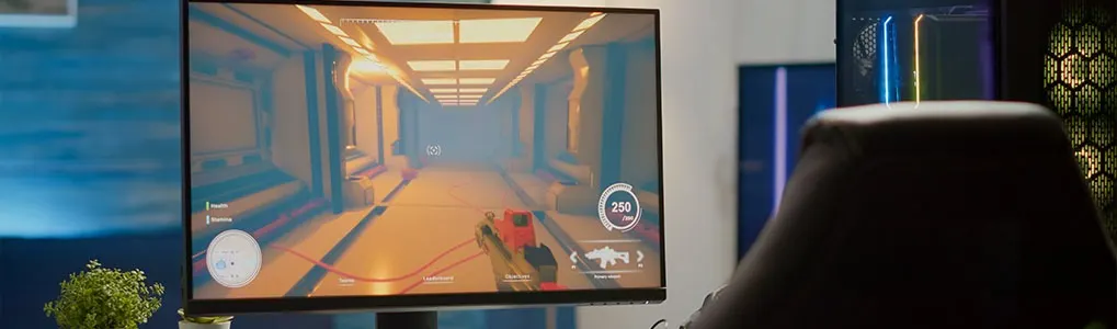 A computer monitor displays a first-person view of a video game, showing a hallway with lights. A chair is in the foreground.