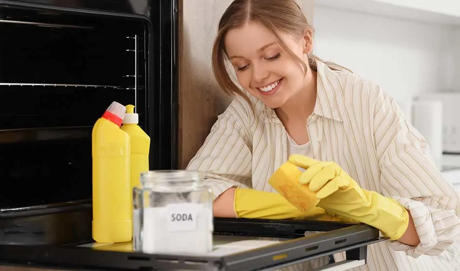 Een glimlachende vrouw met gele handschoenen maakt een oven schoon, met soda en schoonmaakmiddelen erbij.