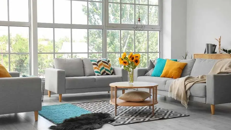 Woonkamer met grijze banken, kleurrijke kussens, een houten tafel met zonnebloemen en een geometrisch vloerkleed.