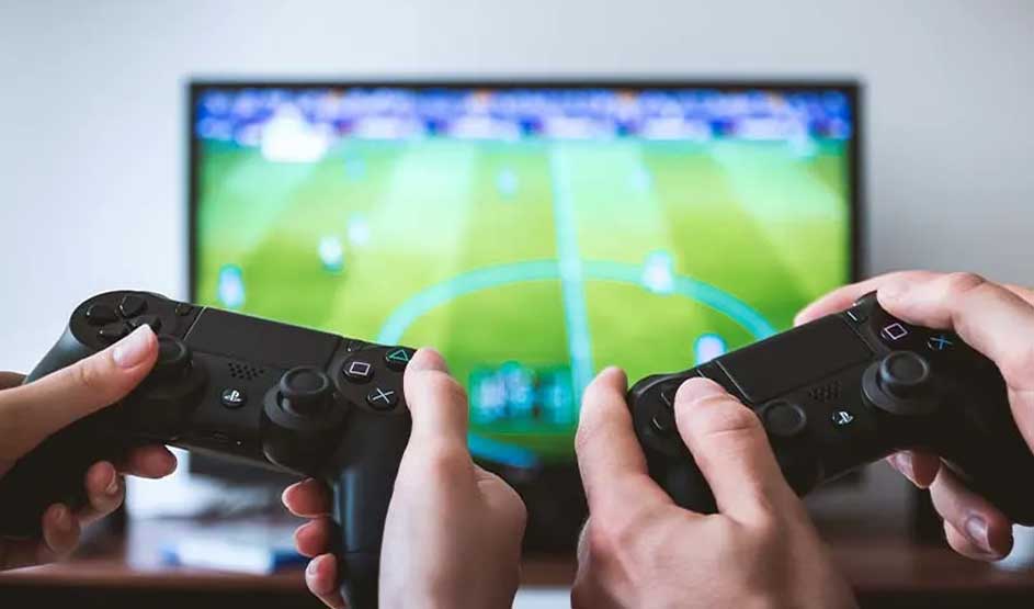 Twee mensen spelen samen een voetbalspel op de PlayStation met twee zwarte DualShock-controllers voor een tv-scherm met voetbalveld.