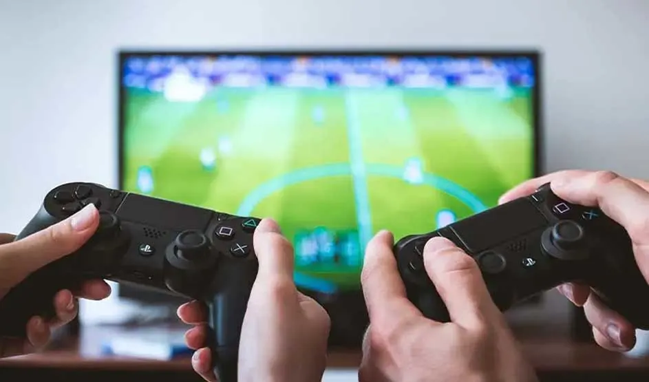 Twee mensen spelen samen een voetbalspel op de PlayStation met twee zwarte DualShock-controllers voor een tv-scherm met voetbalveld.