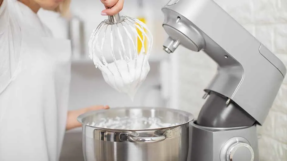 Een zilveren keukenmachine met een metalen kom en een garde vol schuim in een heldere keuken.