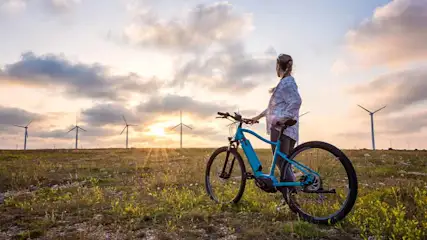 Vrouw met blauwe fiets op een veld met windmolens bij zonsondergang onder een bewolkte hemel.