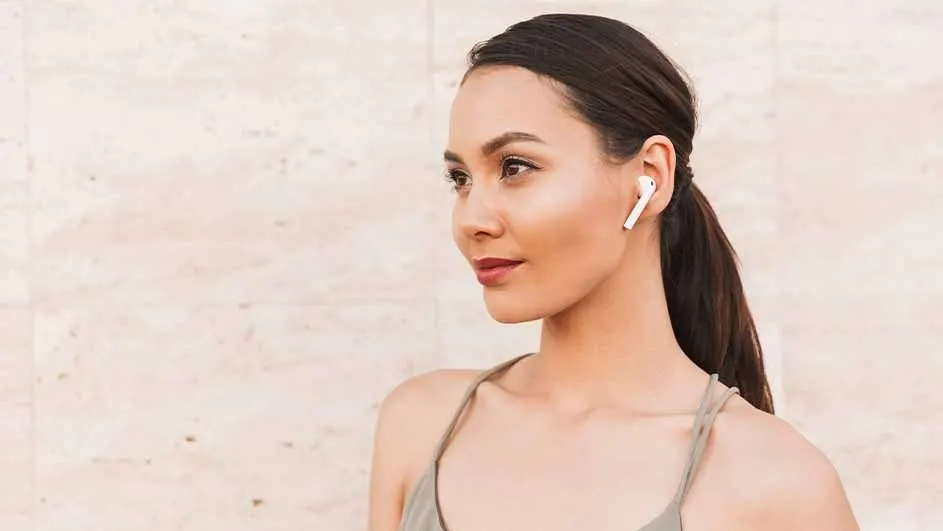 A person with dark hair in a ponytail wears a white earbud. The person wears a light-colored top against a textured, light background.