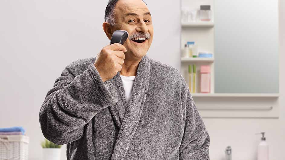 Een man in grijze badjas lacht en scheert zijn snor met een elektrisch scheerapparaat in een badkamer.