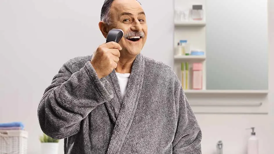 Een man in grijze badjas lacht en scheert zijn snor met een elektrisch scheerapparaat in een badkamer.