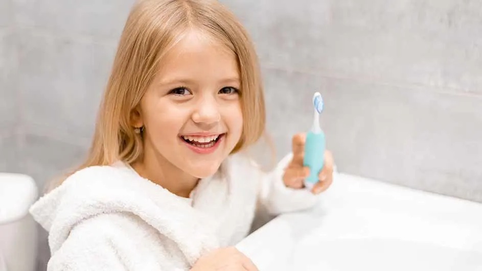 Een kind in een witte badjas zit bij de wastafel en houdt een lichtblauwe elektrische tandenborstel vast in de badkamer.