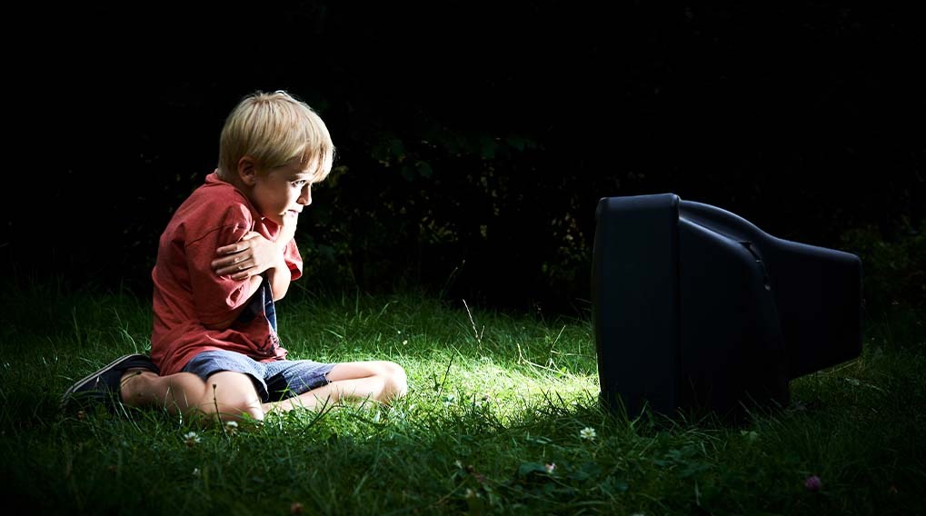 A child in a red shirt and blue shorts sits on green grass, watching a black television in a dark, outdoor setting.