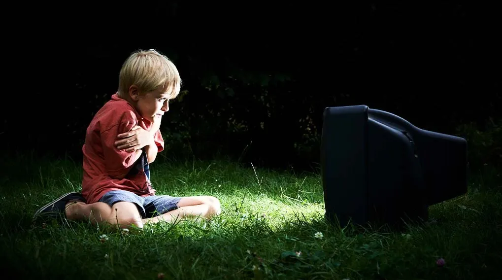 A child in a red shirt and blue shorts sits on green grass, watching a black television in a dark, outdoor setting.