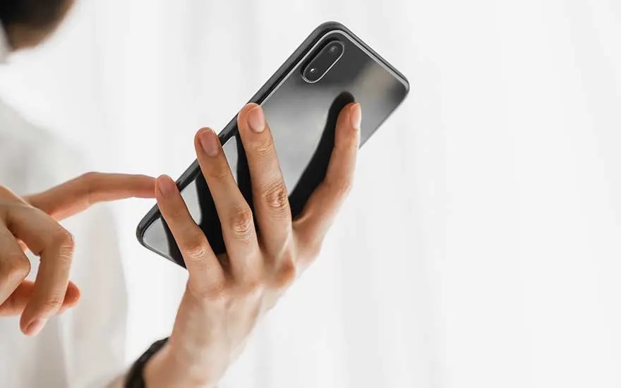 A hand holds a sleek black smartphone upright, fingers curved around the back while the other hand taps lightly against the bright unseen screen.