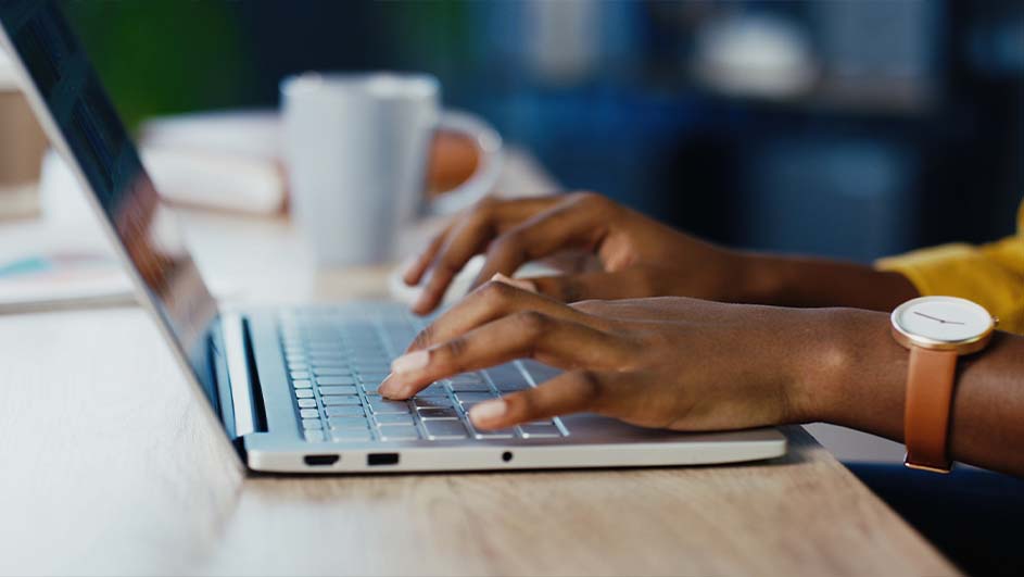Iemand typt op een laptop op een houten tafel met een witte mok op de achtergrond. De persoon draagt een horloge.