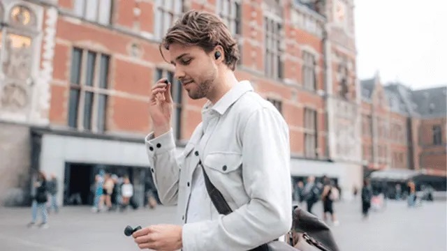 Young man crossing a square with wireless earbuds, holding the case and adjusting one earbud by a historic building.