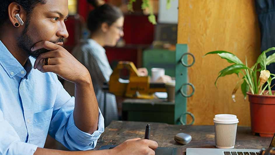 Man in blue shirt uses pen on tablet. Laptop, coffee cup, and potted plant on brown desk. Woman and objects blurred behind.