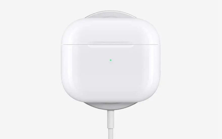 A glossy white AirPods charging case stands upright, connected by a cable, with a tiny green status light glowing on the front.
