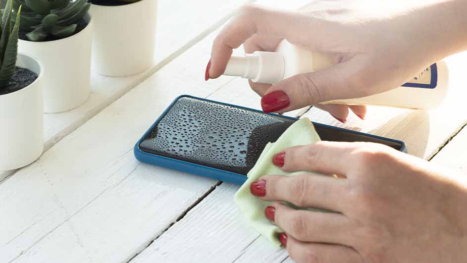 Een telefoon met blauwe rand wordt schoongemaakt met een doek en een spray op een witte houten tafel.
