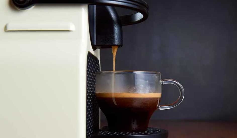 Coffee pours from a cream-colored machine into a clear glass mug. The mug sits on a black drip tray. A dark background.