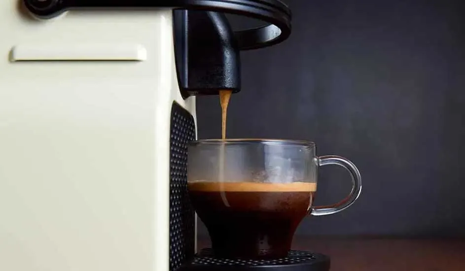 Coffee pours from a cream-colored machine into a clear glass mug. The mug sits on a black drip tray. A dark background.