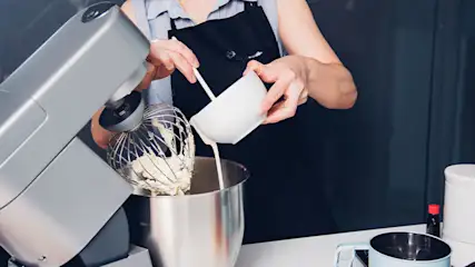 Een persoon giet een vloeistof uit een witte kom in een zilverkleurige keukenmachine met een klopper in een metalen kom.