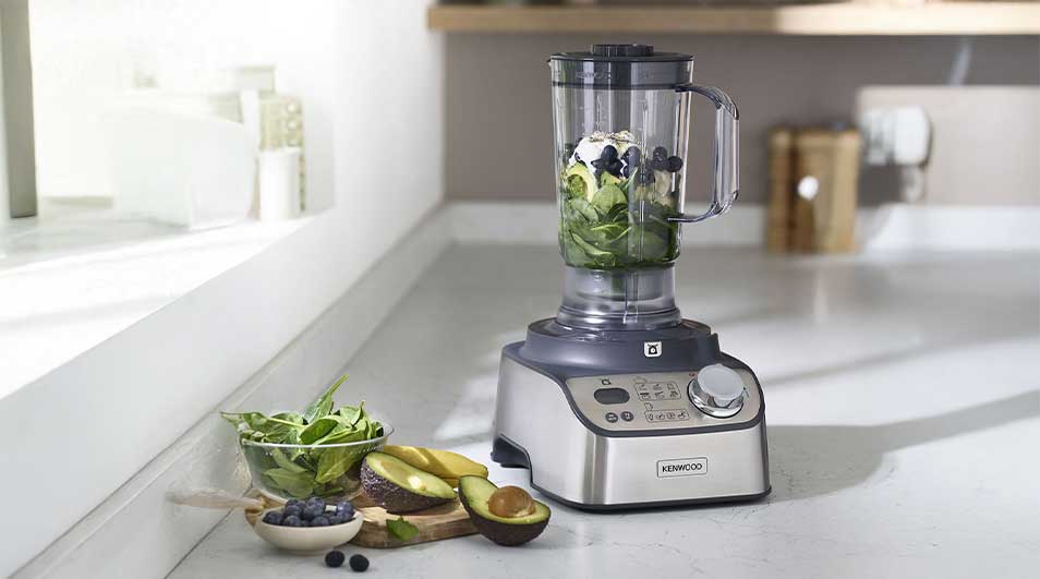 Een krachtige blender met metalen behuizing staat op een aanrecht; in de kan liggen spinazie, avocado, bosbessen en ijsblokjes voor een gezonde smoothie.